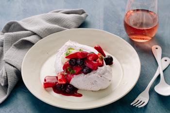 Pavlova dessert dish, surrounded by a grey tea-towel, plastic cutlery and a red drink.