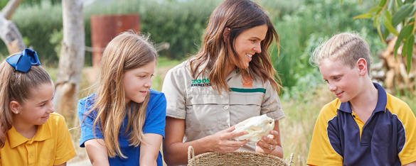 A Zoo keeper sits with a group of three young children dressed in school uniform, as they inspect a basket containing some animal bones.