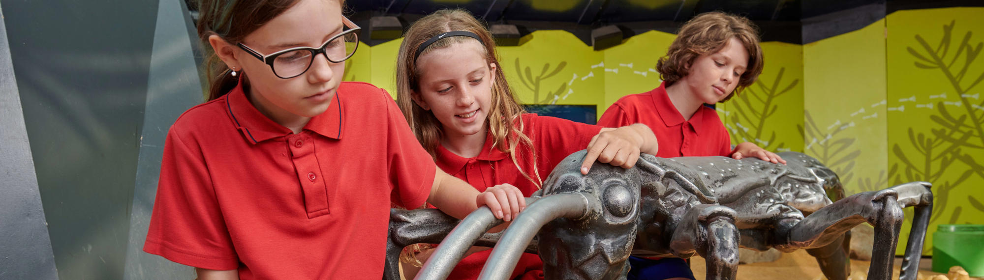 Three students kneeling next to the Lord Howe Island Stick Insect Statue, outside the Butterfly House.