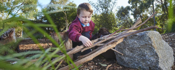 A young school student is crouched on the ground stacking some sticks