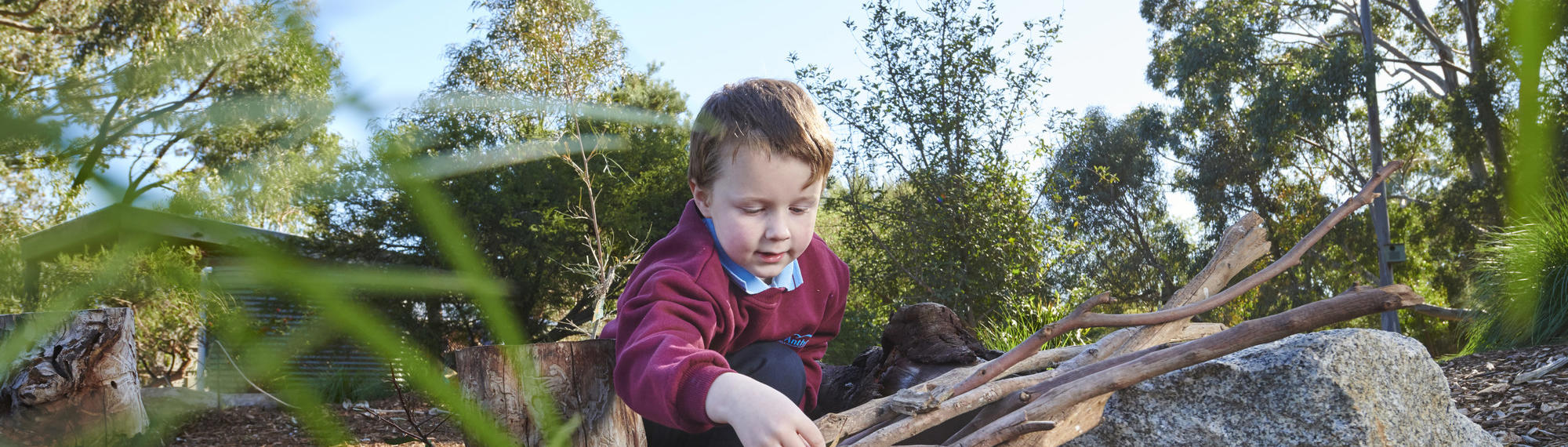 A young school student is crouched on the ground stacking some sticks.