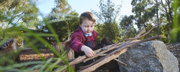 A young school student is crouched on the ground stacking some sticks.
