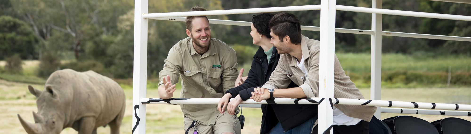 A Keeper stands on the back of a vehicle talking to two people, while a rhino can be seen in the background, all facing left..