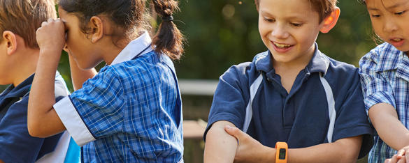 A group of young, primary school aged children sit and play, all dressed in blue uniforms and two wearing orange-banded watches.