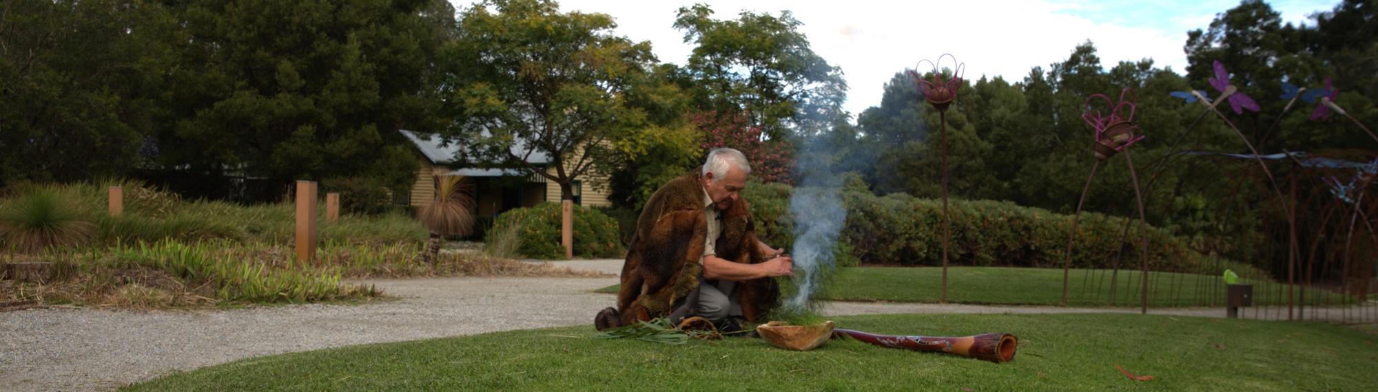 Elder Murrindindi taking part in a Cleansing Ceremony, with a burning grass pile and a didgeridoo.