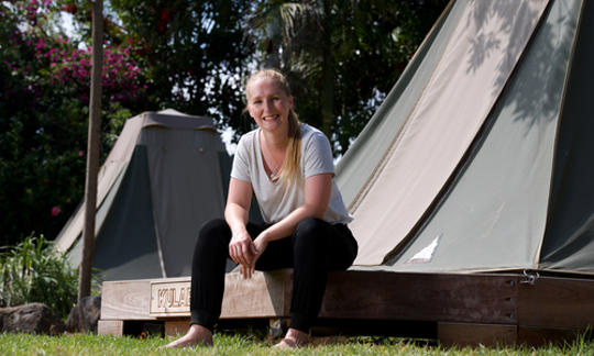 A female with blonde hair is sitting on a wooden platform with a tent in the background. She is smiling at the camera. 
