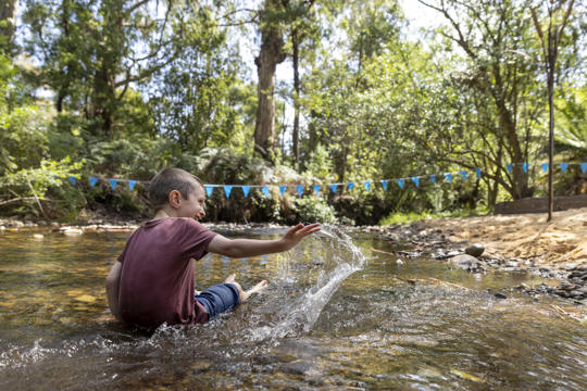 A young guest joyfully slinging water in the shallow Poorneet creek, turned away from the camera.