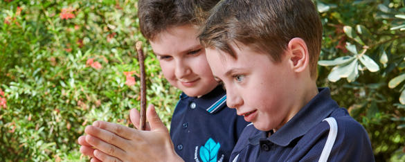 A boy is school uniform rubs a stick between his has as another student looks on