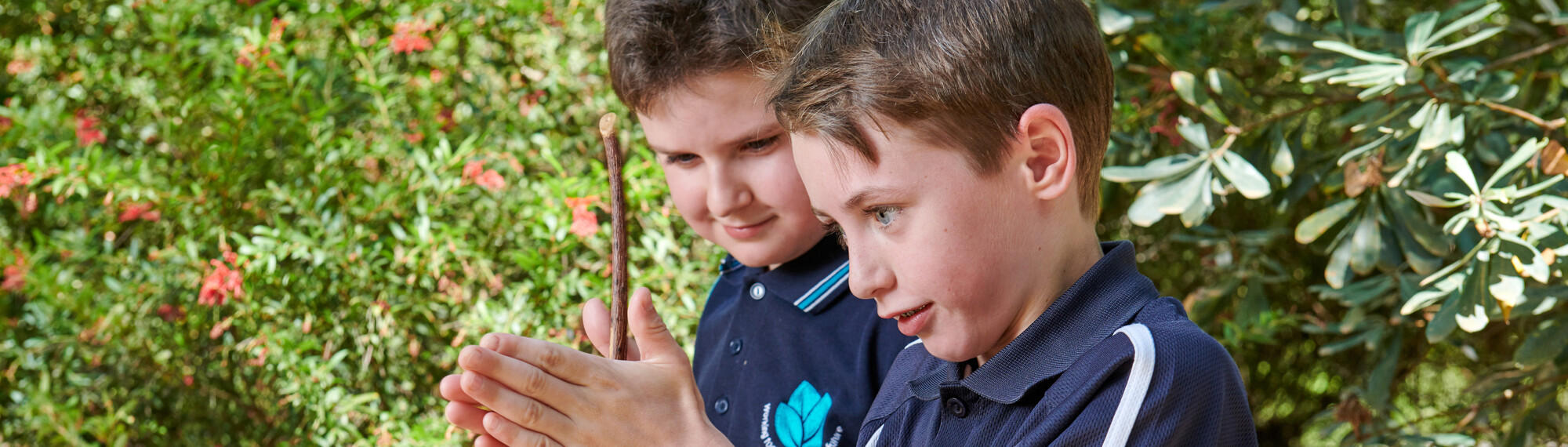 A boy is school uniform rubs a stick between his hands as another student looks on.
