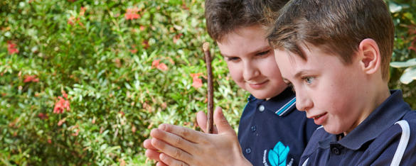 A boy is school uniform rubs a stick between his hands as another student looks on.