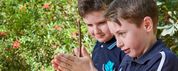 A boy is school uniform rubs a stick between his hands as another student looks on.