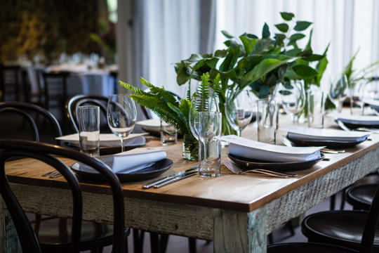 Close-up photo of a wooden table set with greenery, glassware and crockery.