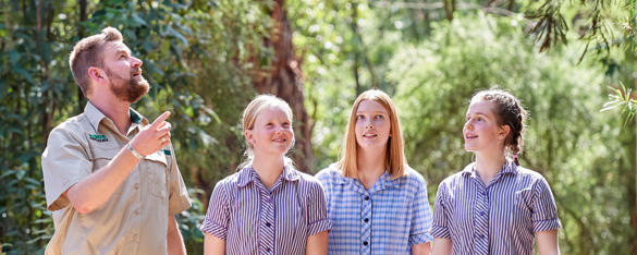 A Zoo staff member points up into the bushland while talking to three school students.