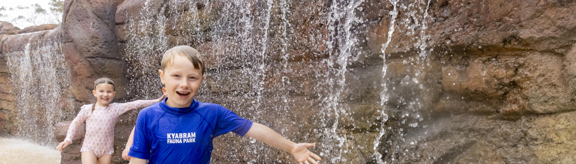 Three young guests run past a small waterfall, running towards and smiling to the camera, with their left arms stretched out to catch water.