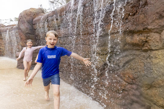 Three young guests run past a small waterfall, running towards and smiling to the camera, with their left arms stretched out to catch water.
