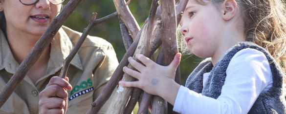 A yound students holds a pile of sticks as she's helped by a zoo staff member