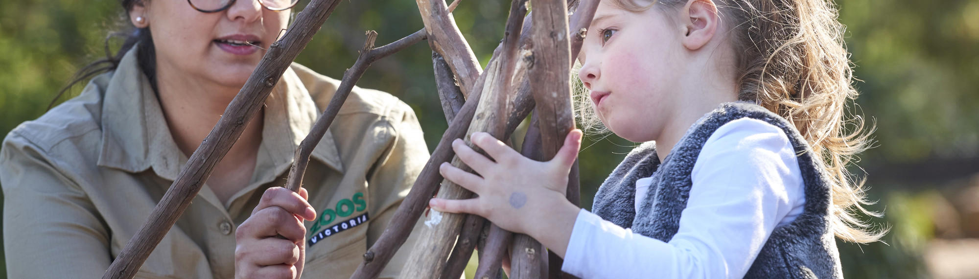 A young student holds a pile of sticks as she's helped by a Zoo staff member.