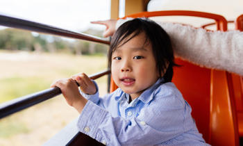 Young boy looking at camera sitting on an orange bus seat holding a bar across the window