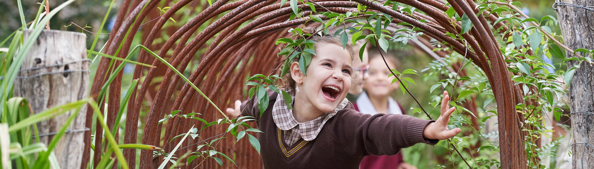 Girl in brown school uniform smiles as she runs through an archway, while two other students can be seen behind her.
