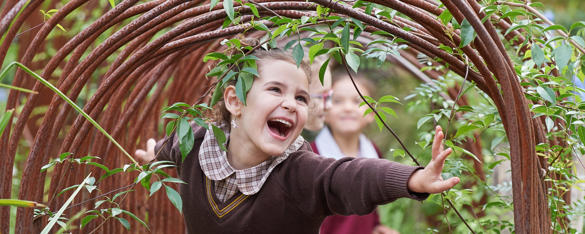 Girl in brown school uniform smiles as she runs through an archway, while two other students can be seen behind her.
