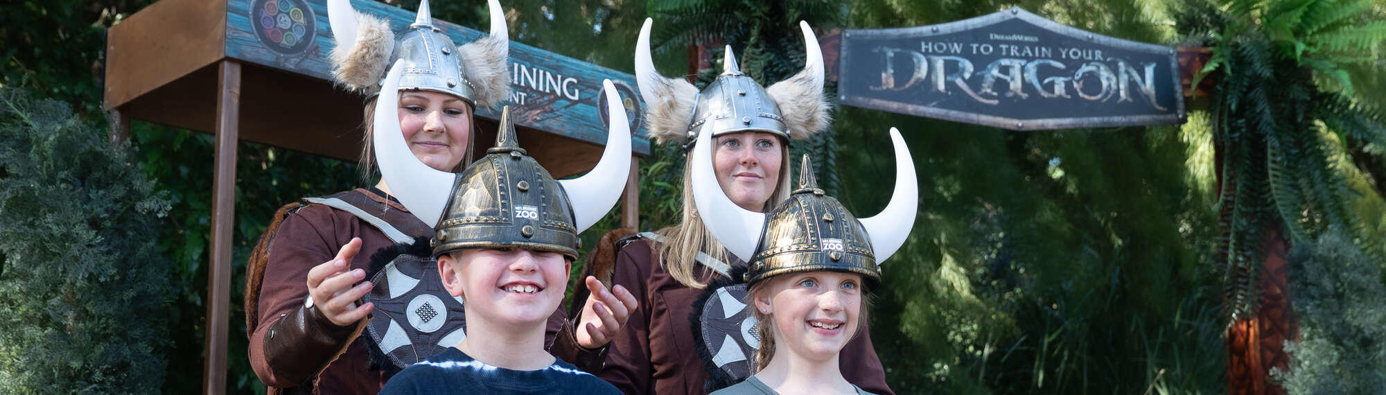 "How To Train Your Dragon"; Two young guests having their helmets put on by two "Vikings," all smiling.
