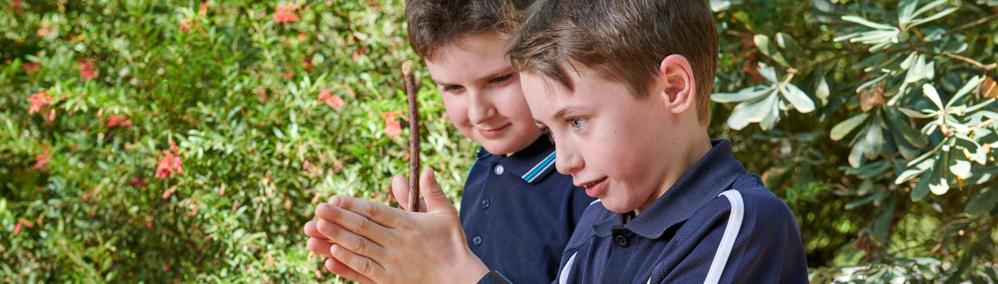 Student rubs a stick between his hands, as another student looks on, both facing left.