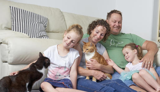 A family of four sitting in front of a couch patting two cats and smiling