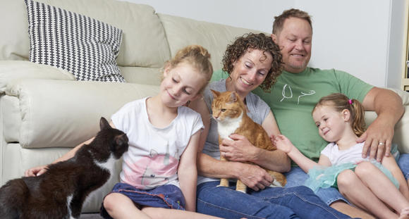 A family of four sitting in front of a couch petting two cats and smiling.