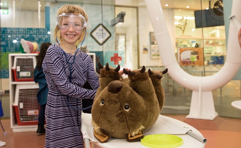 A kid wearing glasses, "examining" a wombat plush at Future Vets, while she smiles right to the camera.
