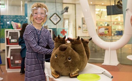 A kid wearing glasses, "examining" a wombat plush at Future Vets, while she smiles right to the camera.