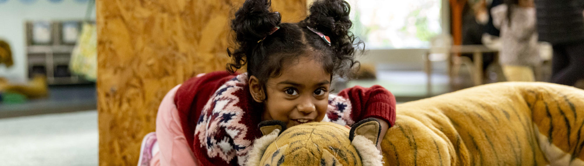 A young guest cuddles the large plush Tiger of Keeper Kids, while she faces the camera, crouching and smiling.