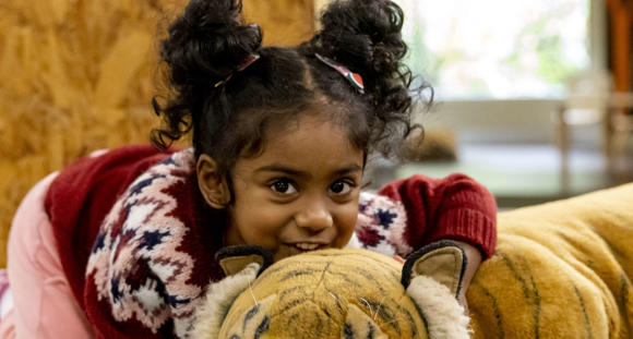 A young guest cuddles the large plush Tiger of Keeper Kids, while she faces the camera, crouching and smiling.