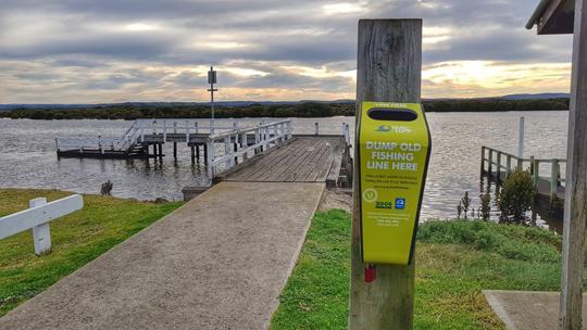 A bin on a wooden pole in front of a jetty over an inland waterway; the bin has text on it that reads "Dump old fishing line here"