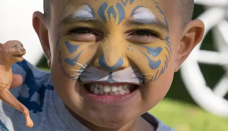 A young guest with tiger face-paint grins to the camera, while holding a lioness toy in his right hand.