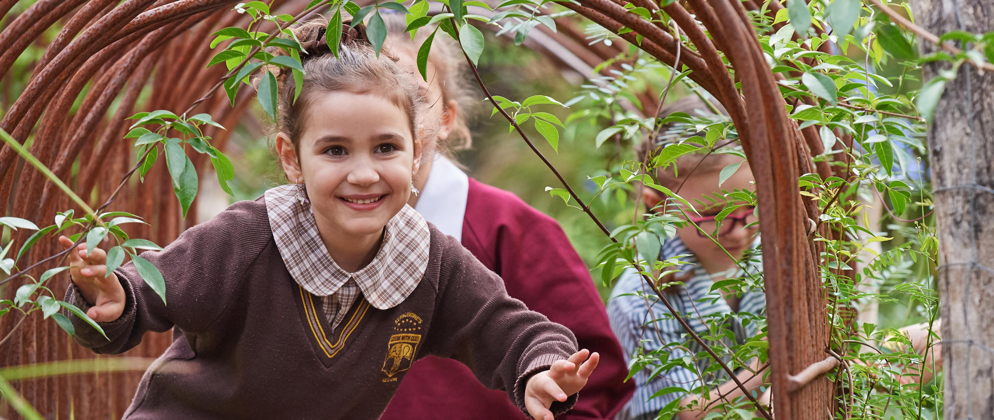 Young student (in brown school uniform) crouching through a forest archway, with another two students behind her.