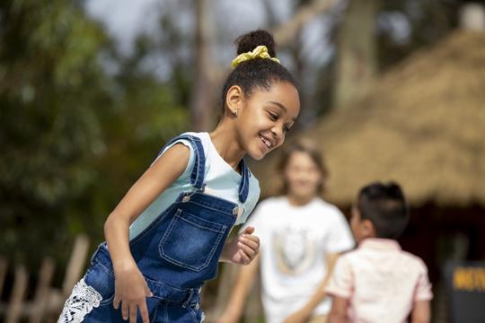 A young girl in overalls is smiling and running around. Other kids are blurred out in background.
