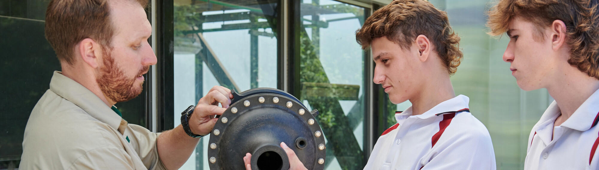 A Zoo employee shows two teen students, dressed in white and red school uniforms, a piece of pumping machinery.