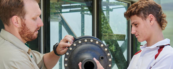 A Zoo employee shows two teen students, dressed in white and red school uniforms, a piece of pumping machinery.