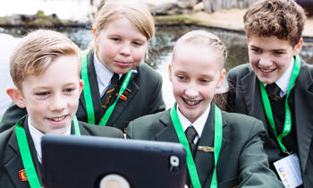 Four students looking at tablet device while smiling, with the Hippo River behind them..