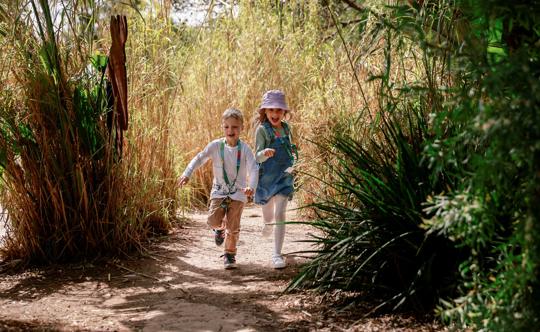 Two children in zoo lanyards are running down a pathway surrounded by long grass and and bamboo.