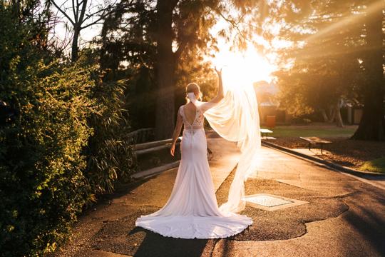 Wedding ceremony at dusk, as the Bride flutters her veil above her right and to the sun, facing away from the camera.