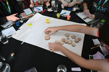 At least five guests gathered around a black table, all engaged in a craft activity with paper, scissors, markers, three tubs of modelling clay and fifteen small wooden discs.