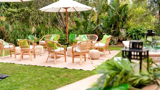 Chairs and umbrella set up on a picnic rug on a green lawn surrounded by lush green bushes