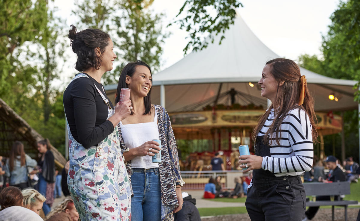 A group of three woman laughing together with drinks, the carousel in the background with dusk lighting