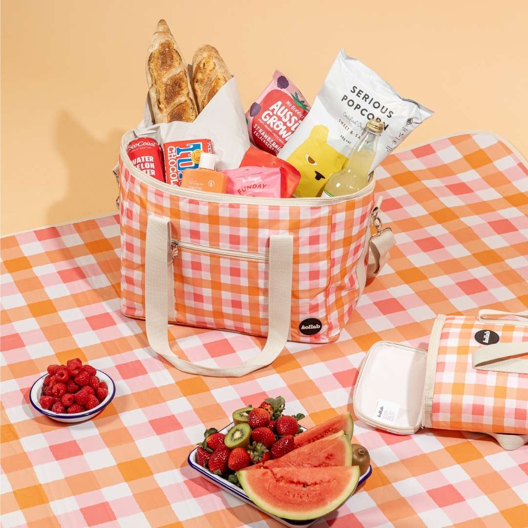 Hamper of bread, berries, flavoured drinks, chocolates and popcorn; two bowls of raspberries, strawberries, watermelon and kiwi fruit; one empty bag and table covering with orange-and-white cross-hatch pattern.