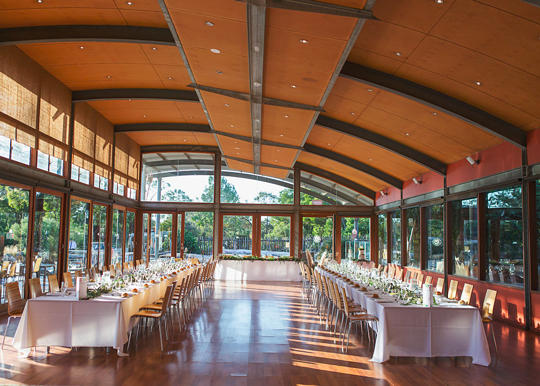 A room with two long tables and chairs lined up on either side of the table. The tables have white table cloths on them and greenery.