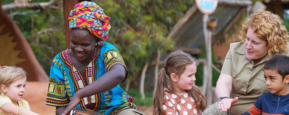 Three young guests sit with two women, one of whom is dressed in traditional colourful clothing and another in a khaki Zoos Victoria uniform.