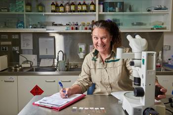 A smiling Vet, dressed in khaki, sits in front of a microscope with a clipboard and pen in her hand.