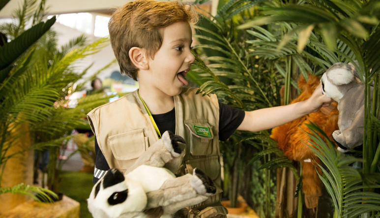 A young guest in a Keeper vest excitedly plays with two plush Lemurs, with an Orangutan and ferns behind him.