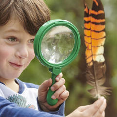 Kinder Keeper Kid examines a brown-and-orange feather with a green-framed magnifying glass.